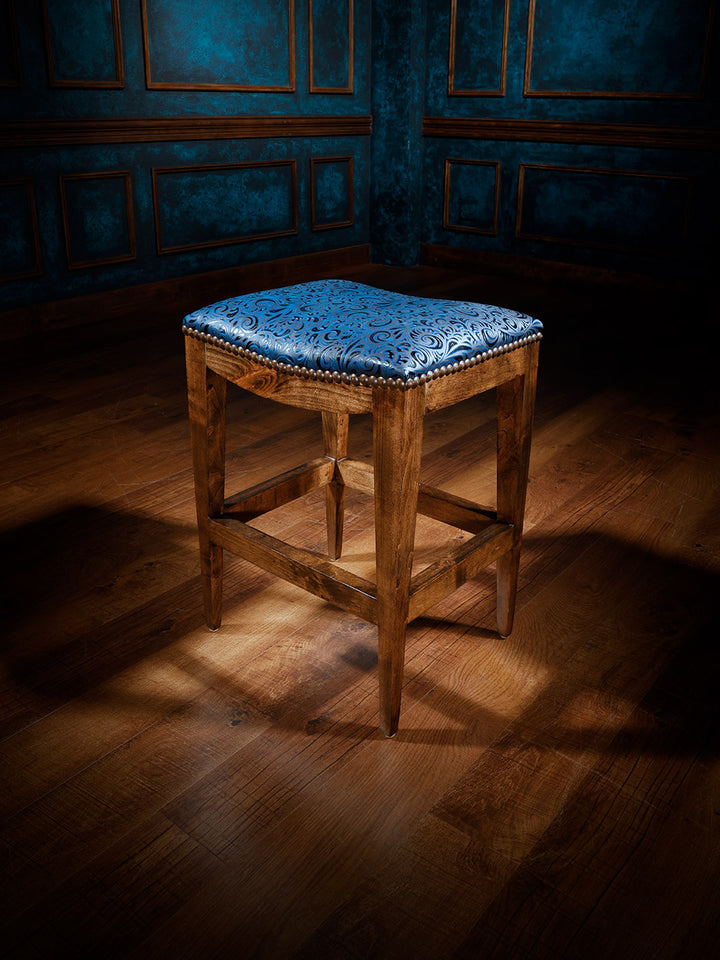 A wooden western saddle stool with a blue upholstered seat, positioned on a hardwood floor against a paneled wall.