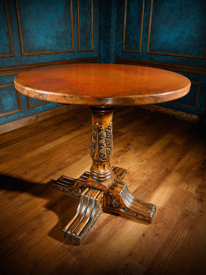 A round pub table with a solid copper top and a wooden, hand-carved pedestal base, placed on a hardwood floor with blue paneling in the background.