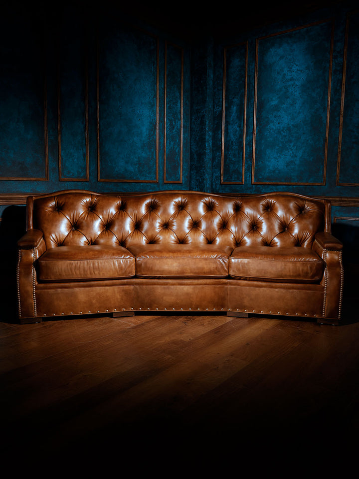 A full view of a curved, brown tufted leather sofa with three seat cushions and nailhead trim, set against a dark blue paneled wall on a hardwood floor. The sofa has a classic Western style with a rich caramel tone and deep button-tufted backrest.