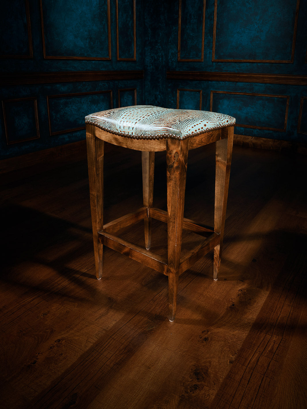 A saddle stool with a wooden frame and a turquoise crocodile patterned seat, placed on a dark wooden floor with a blue paneled wall in the background.