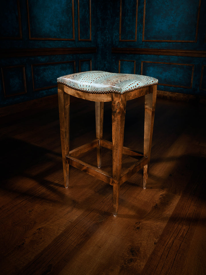 A saddle stool with a wooden frame and a turquoise crocodile patterned seat, placed on a dark wooden floor with a blue paneled wall in the background.