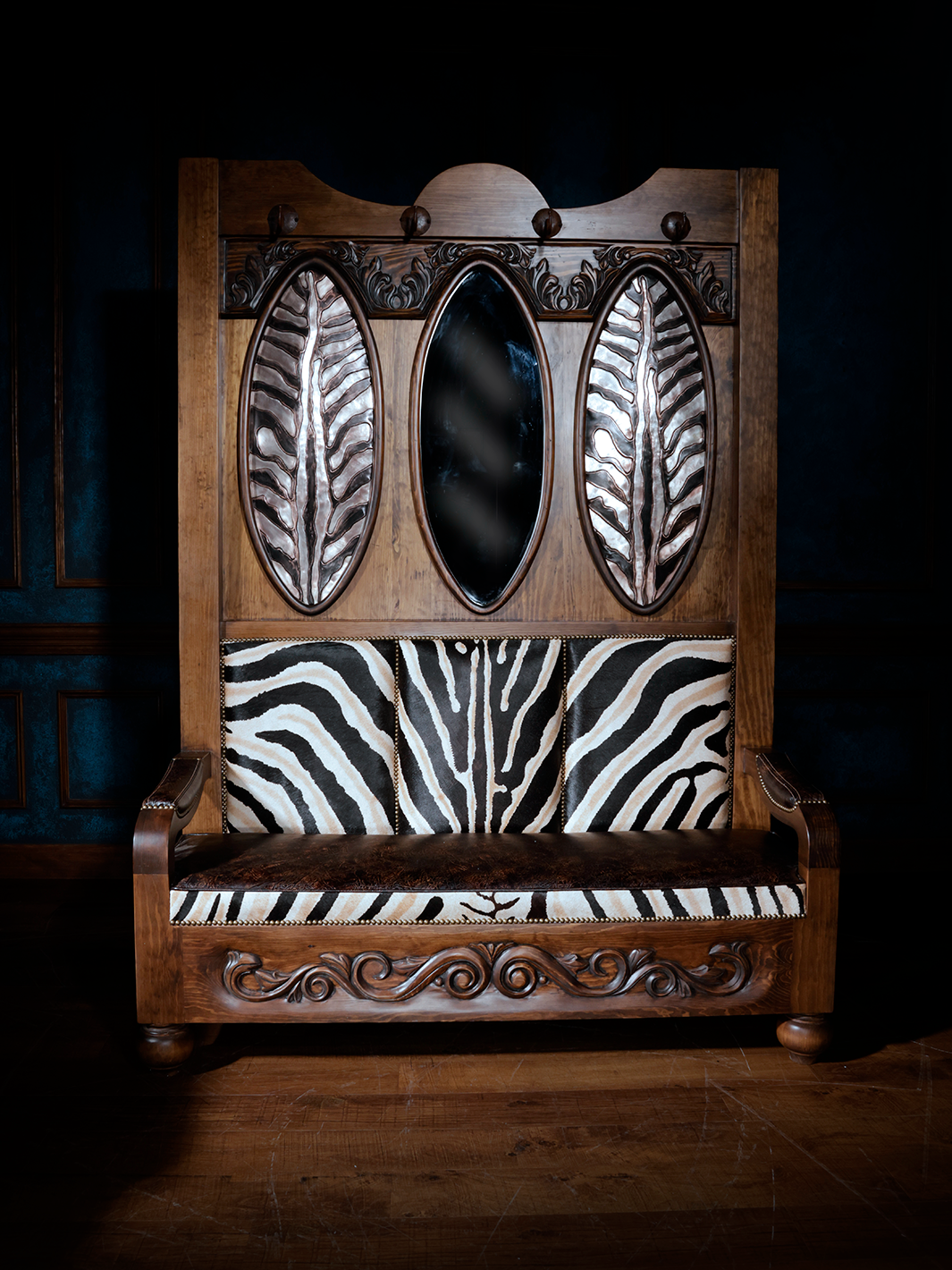 A decorative wooden coat rack with a zebra print leather upholstered seat, featuring iron conchos with hooks, and carved floral and scroll motifs.
