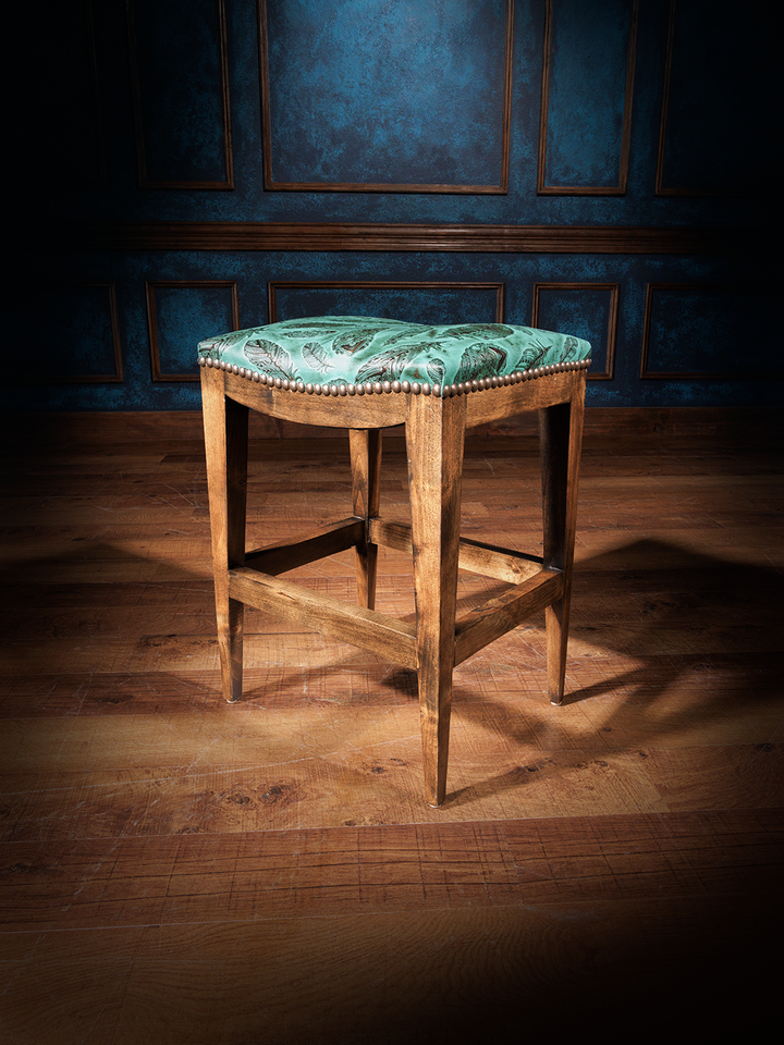 A handcrafted leather saddle stool with a green floral pattern on the seat, a brown wooden frame, and a tufted backrest.