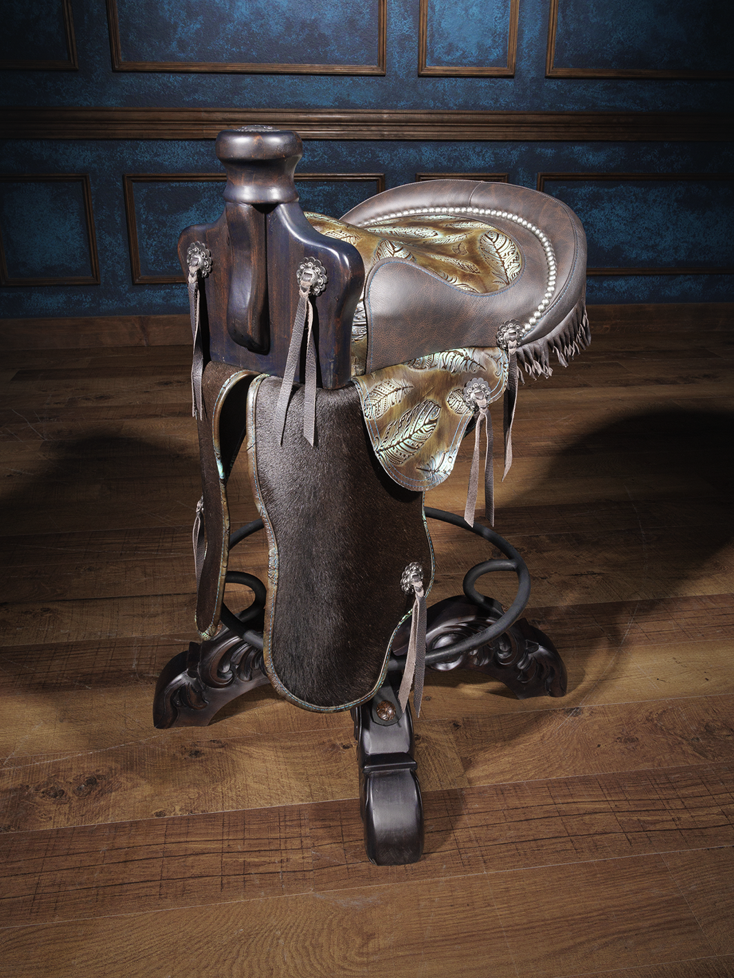 A saddle bar stool with brown full grain leather, silver finished conchos, and turquoise colored genuine leather with feather embossing. It features a round, hand-forged iron medallion embossed with a floral pattern, and is set on a hand-turned wood trumpet pedestal with four hand-carved wood feet.