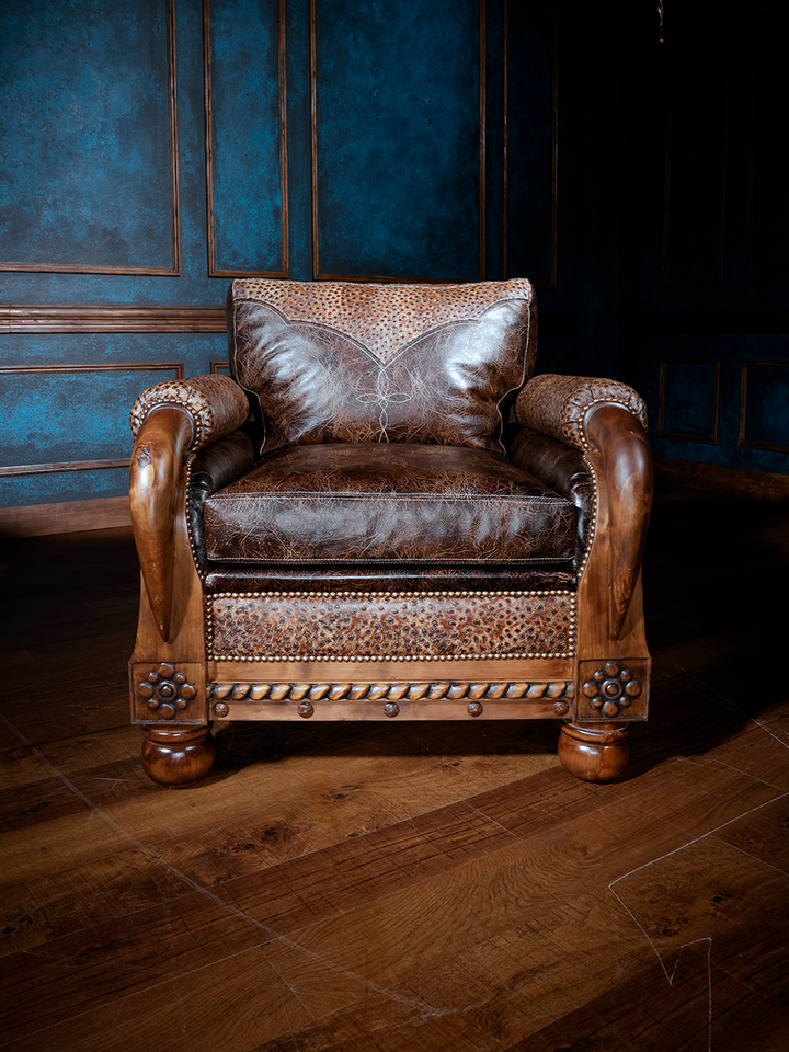 Front view of a luxurious western accent chair with distressed leather seating, decorative nailhead trim, and tooled leather along the front apron. Intricately carved rosettes and turned legs complete this high-end ranch aesthetic.