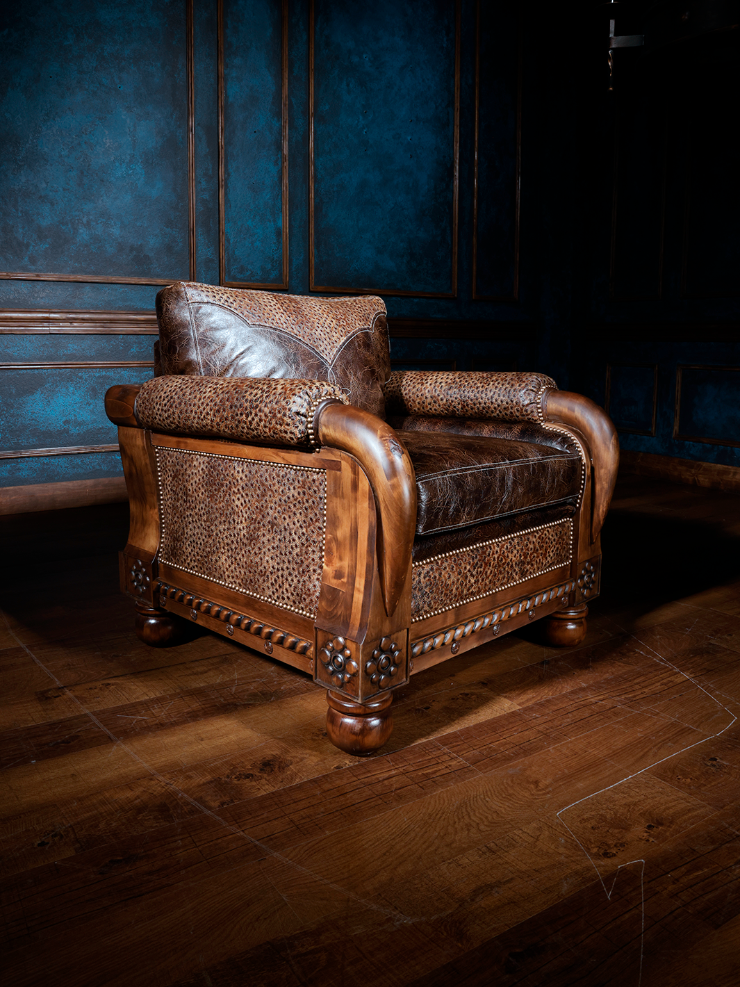 Carved Western armchair featuring rich brown leather cushions, embossed leather accents, and a hand-carved wood frame with floral detailing and curved scroll arms. A rugged yet refined piece of heirloom-quality seating.