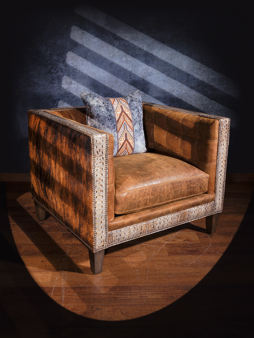 Western box-style leather chair with a structured square silhouette, featuring hair-on-hide side panels, embossed leather trim, and a distressed leather cushion. A patterned accent pillow adds a bold southwestern touch.
