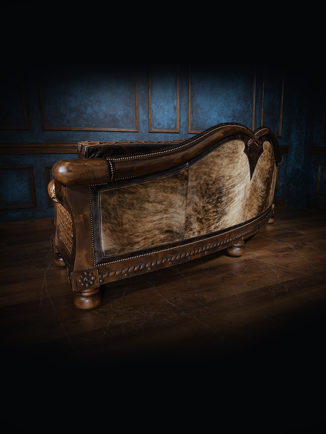 Rear view of a handcrafted western leather sofa showcasing a curved back upholstered in light brown cowhide with nailhead trim and carved wood accents.