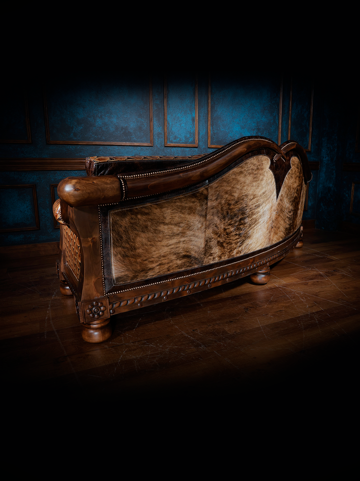 Rear view of a handcrafted western leather sofa showcasing a curved back upholstered in light brown cowhide with nailhead trim and carved wood accents.
