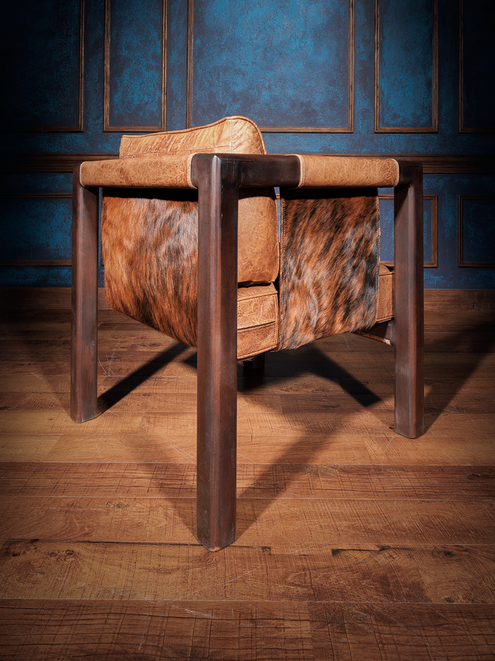 Western Santa Fe sling chair with a rich wooden frame and panels of warm distressed leather and brindle cowhide. The chair’s back and seat sling subtly into the frame, offering a blend of southwestern charm and contemporary form.
