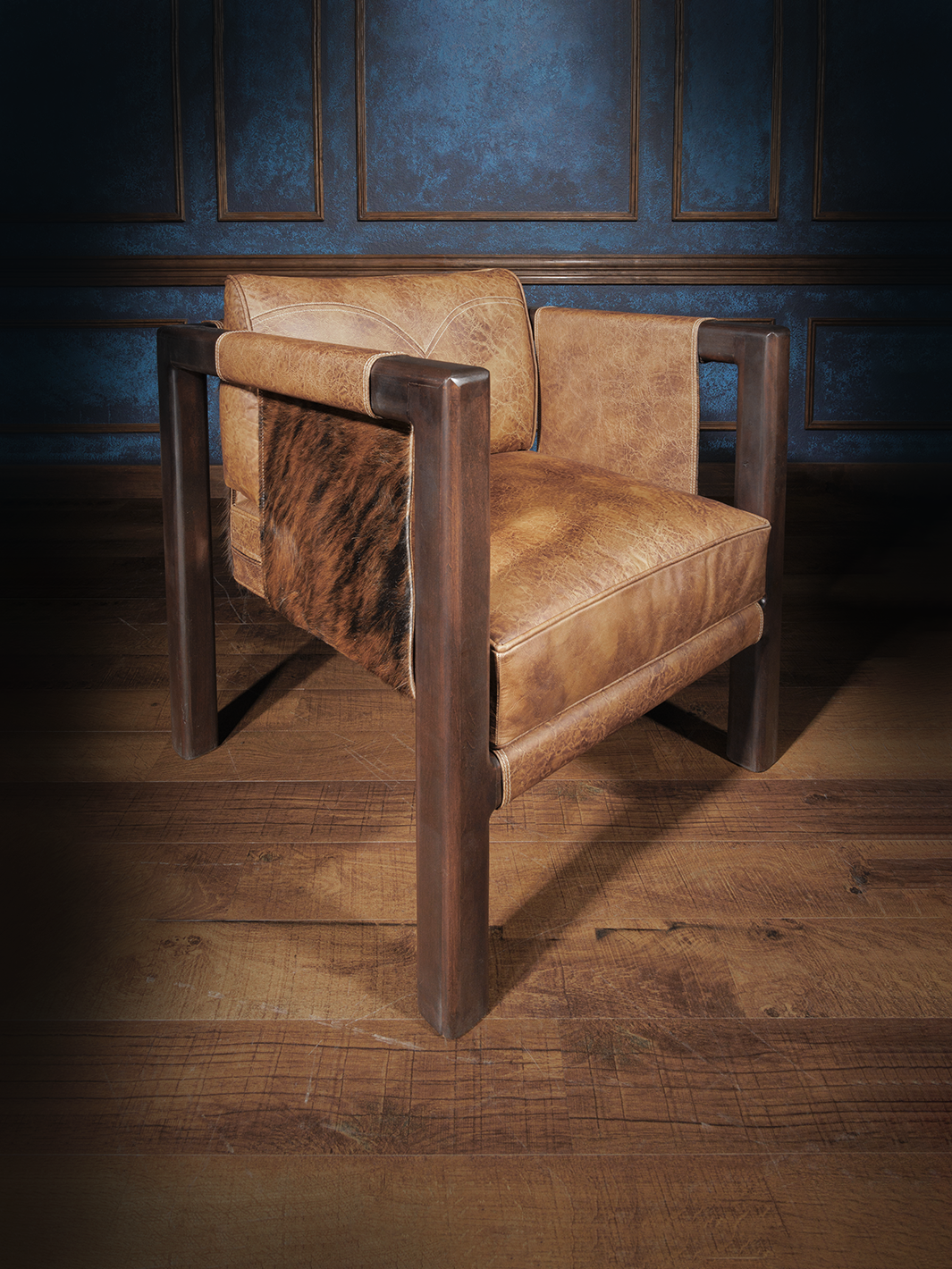 Angled side view of a rustic leather and hide accent chair, featuring tubular wood legs, natural hide sides, and curved topstitching on the backrest. The piece offers bold geometry softened by organic textures.