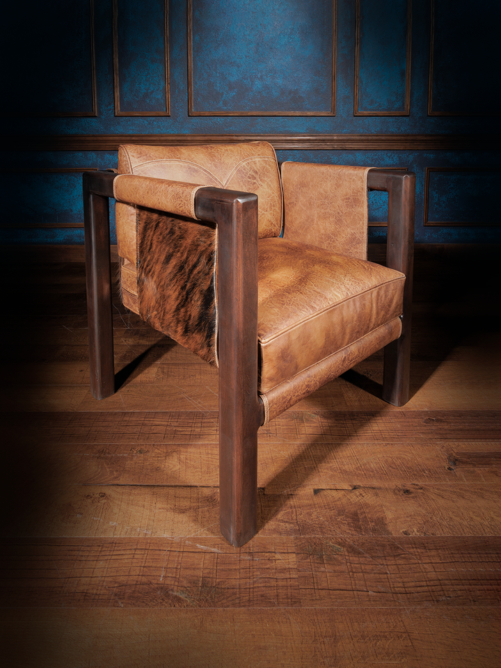 Angled side view of a rustic leather and hide accent chair, featuring tubular wood legs, natural hide sides, and curved topstitching on the backrest. The piece offers bold geometry softened by organic textures.