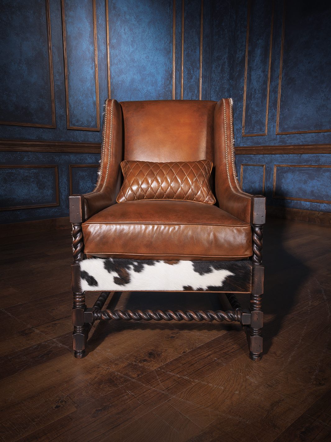 Western cowhide and leather accent chair with a high back, saddle-toned leather seat and backrest, and dark carved wood legs. A quilted leather lumbar pillow adds refined comfort to its rugged silhouette.