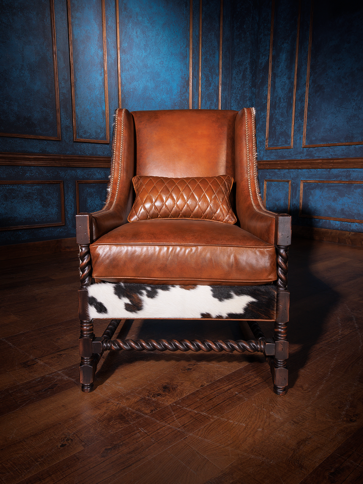 Western cowhide and leather accent chair with a high back, saddle-toned leather seat and backrest, and dark carved wood legs. A quilted leather lumbar pillow adds refined comfort to its rugged silhouette.