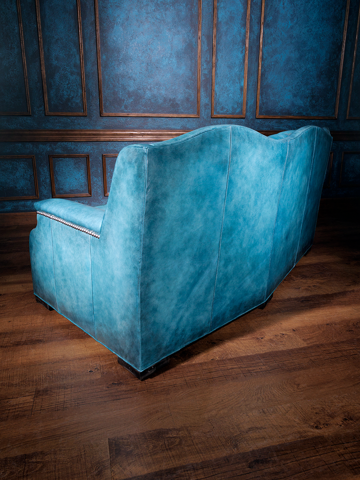 Back view of a bright blue western leather couch featuring a high, scalloped back and smooth paneling on hardwood floors.