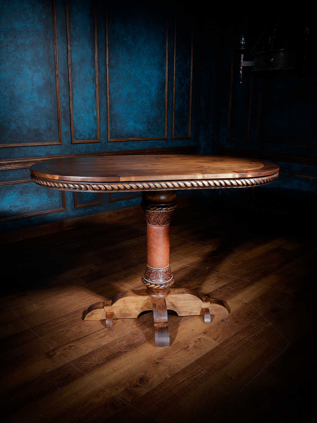 A circular wooden pub table with intricate rope trim carvings and a decorative pedestal base, placed on a wood floor with a blue-painted wall in the background.