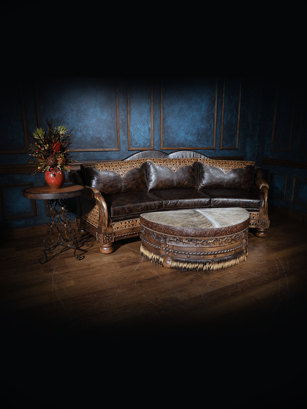Western leather sofa with dark brown cushions and an intricately carved wood frame, paired with a round cowhide ottoman and rustic accent table.