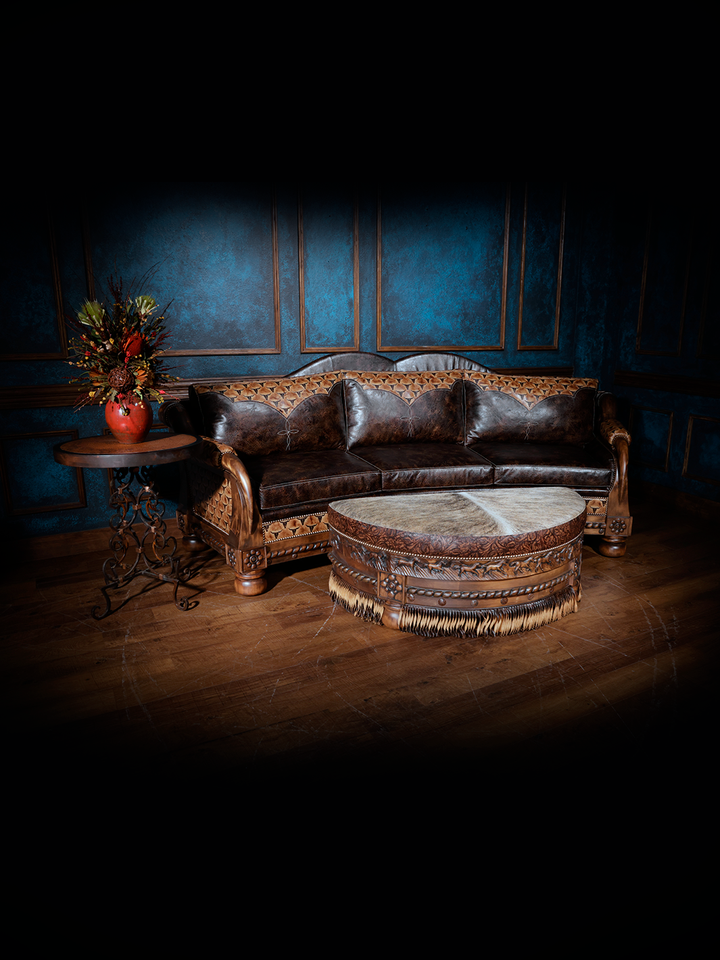 Western leather sofa with dark brown cushions and an intricately carved wood frame, paired with a round cowhide ottoman and rustic accent table.
