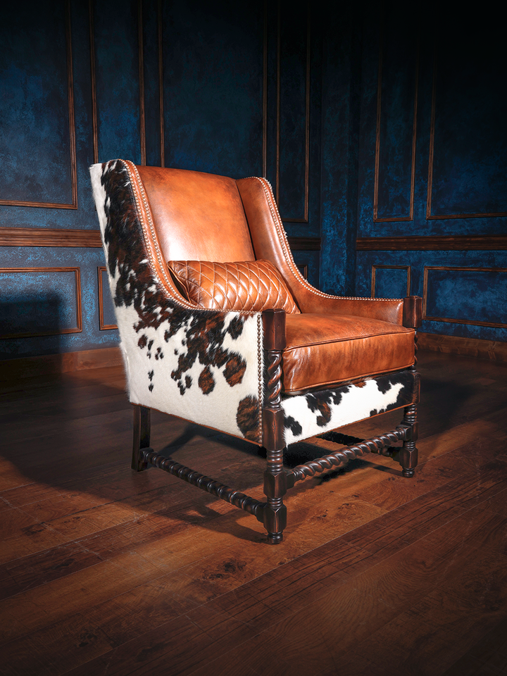 Side view of a rustic western accent chair showcasing smooth leather paired with dramatic black and white cowhide upholstery. Nailhead trim and barley twist legs complete the elevated ranch aesthetic.