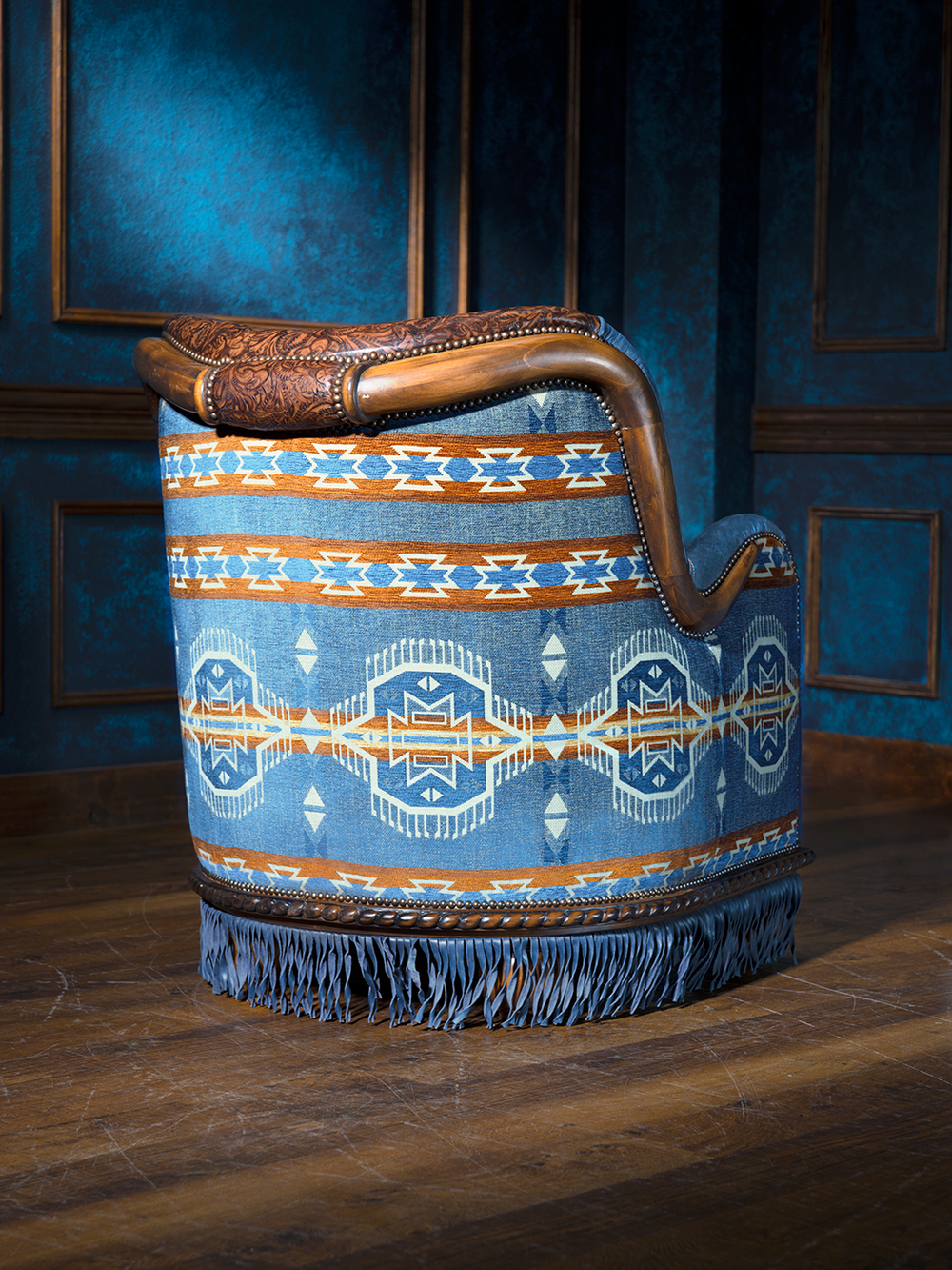 Back view of a western-style chair upholstered in southwestern patterned fabric with tones of blue, rust, and cream. A carved wood top rail and leather fringe complete the high-end ranch look.