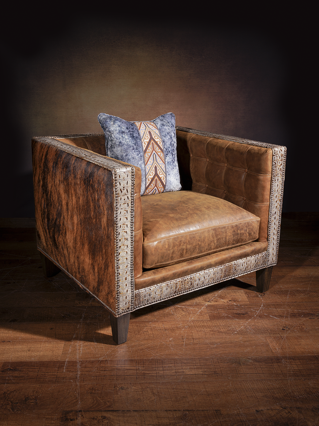 Rustic boxy leather armchair upholstered with smooth caramel leather, trimmed in tooled leather, and flanked by rich cowhide panels. Styled with a textured blue and rust-patterned throw pillow for western flair.