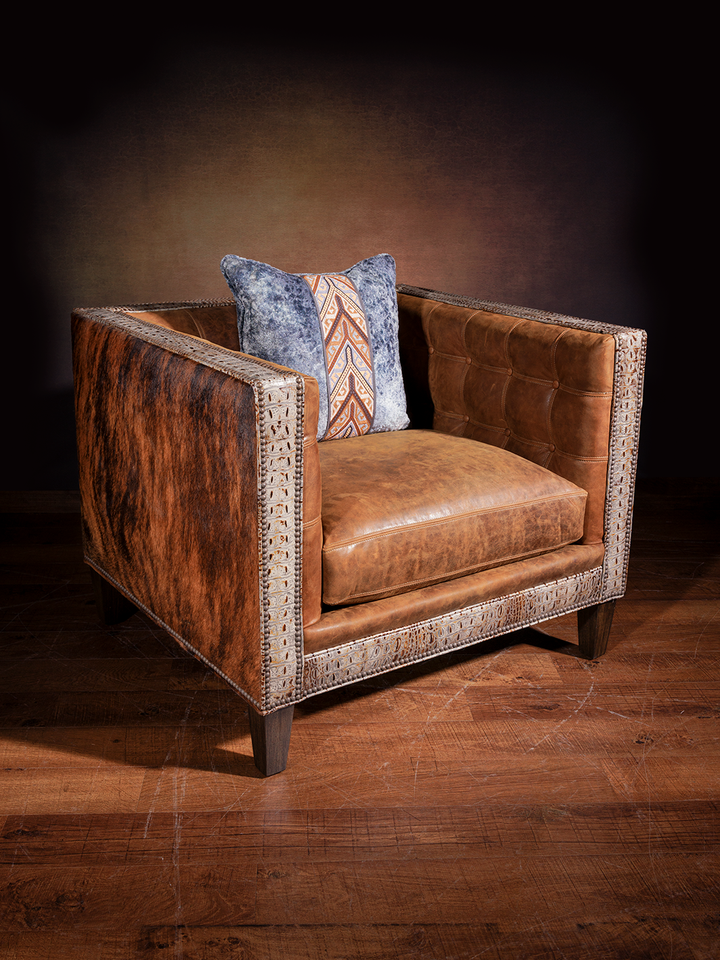 Rustic boxy leather armchair upholstered with smooth caramel leather, trimmed in tooled leather, and flanked by rich cowhide panels. Styled with a textured blue and rust-patterned throw pillow for western flair.