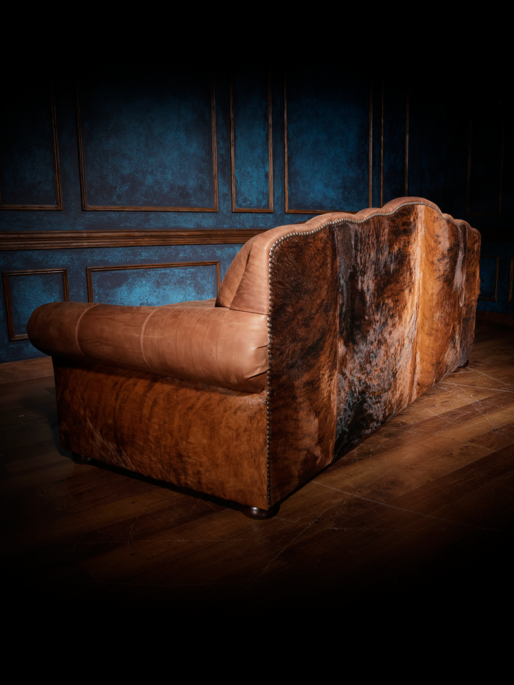 Back view of a rustic leather and cowhide sofa featuring a scalloped top, rolled arms, and richly textured hide upholstery.