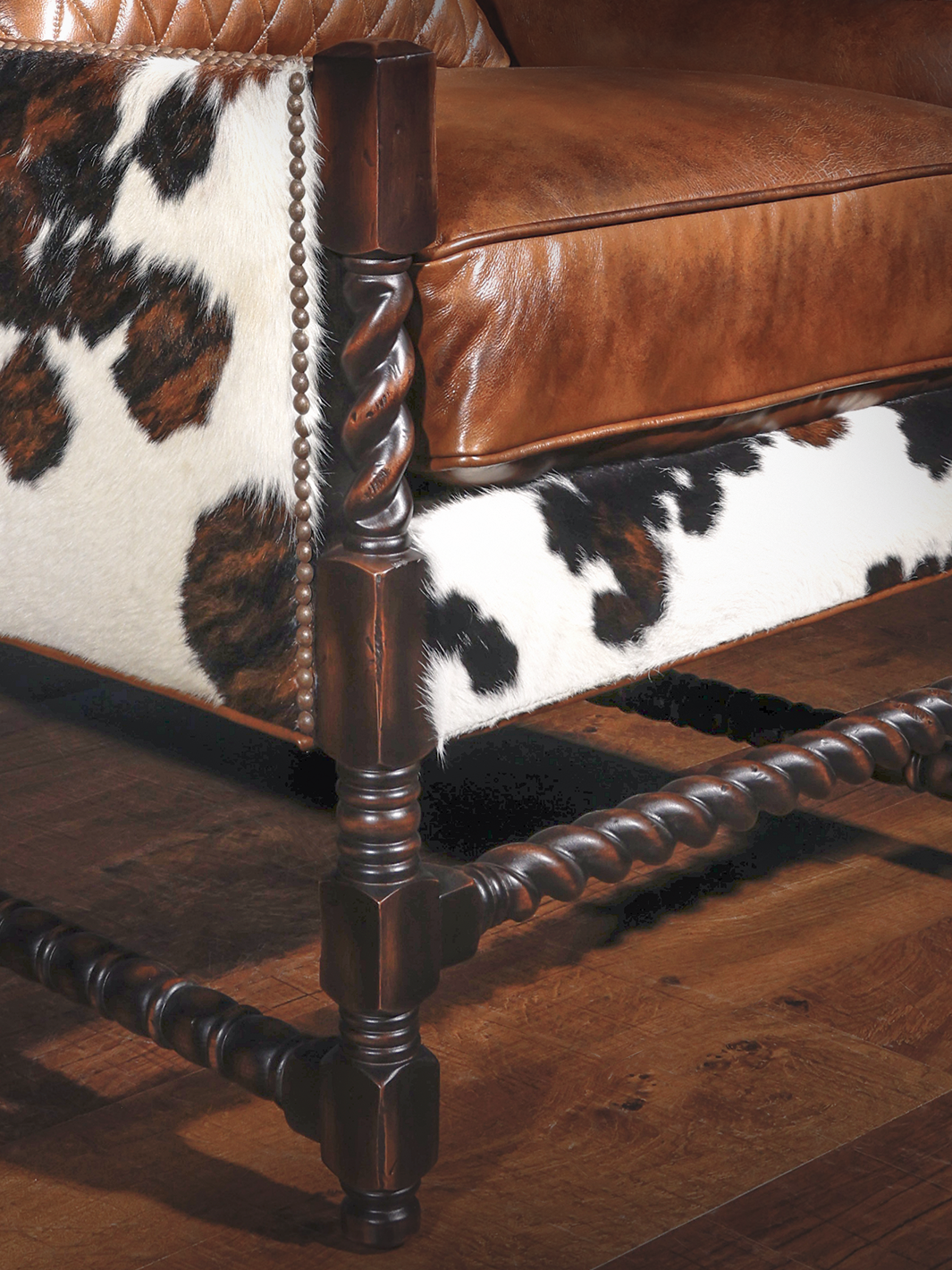 Close-up of the chair's lower corner, highlighting the dark walnut barley twist legs, decorative nailhead detailing, and natural cowhide upholstery wrapping the base.
