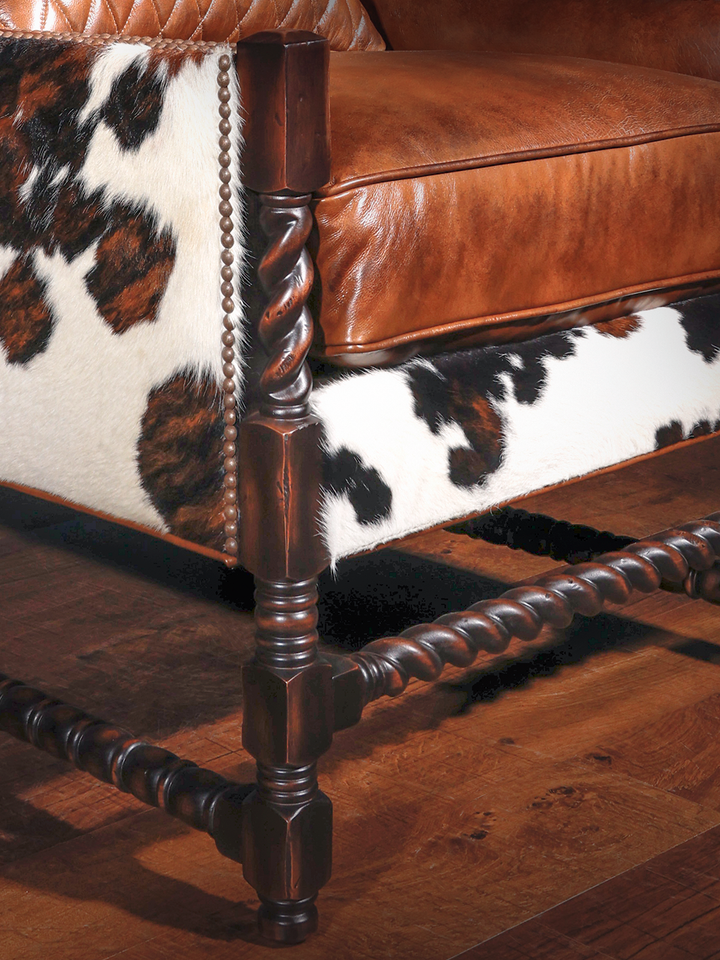 Close-up of the chair's lower corner, highlighting the dark walnut barley twist legs, decorative nailhead detailing, and natural cowhide upholstery wrapping the base.