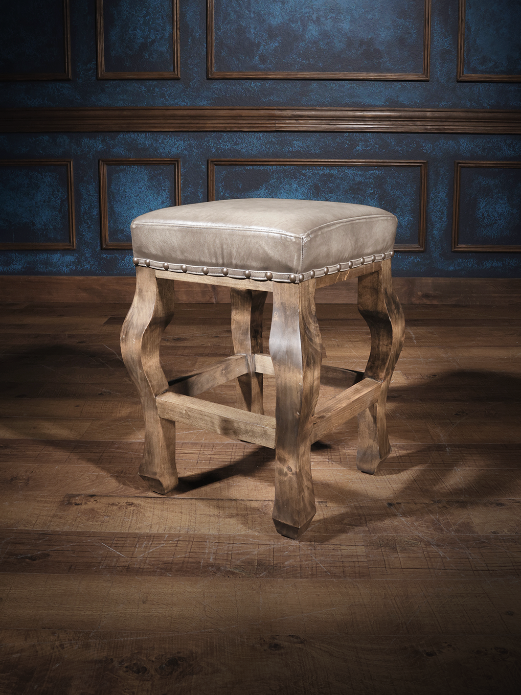 A Western Sterling Leather Saddle Stool with a beige seat and brown wooden legs, placed against a blue panelled wall.