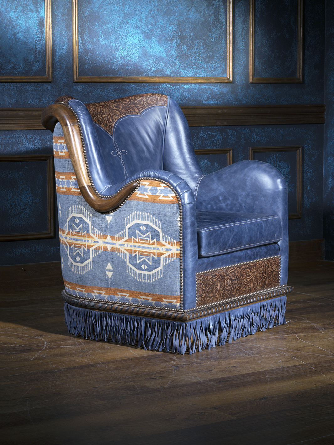 Angled view of a bold western accent chair featuring blue leather, tribal-inspired upholstery on the back and sides, and intricate floral tooled leather panels. The chair is finished with nailhead trim and floor-grazing fringe for added flair.