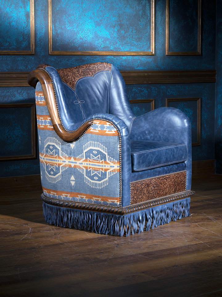 Angled view of a bold western accent chair featuring blue leather, tribal-inspired upholstery on the back and sides, and intricate floral tooled leather panels. The chair is finished with nailhead trim and floor-grazing fringe for added flair.