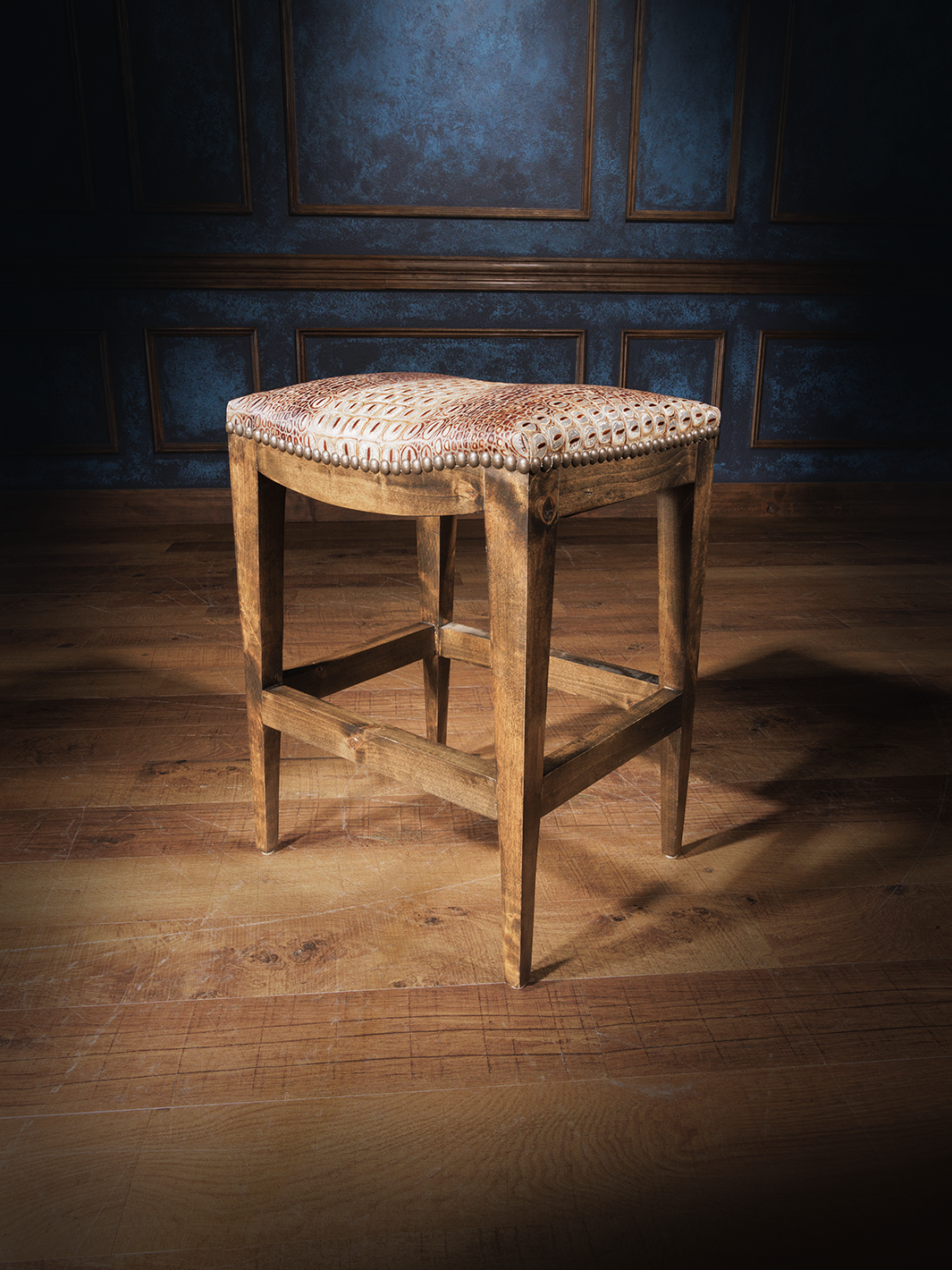 A brown crocodile leather saddle stool with a patterned fabric seat cushion, placed on a hardwood floor with a blue panelled wall in the background.
