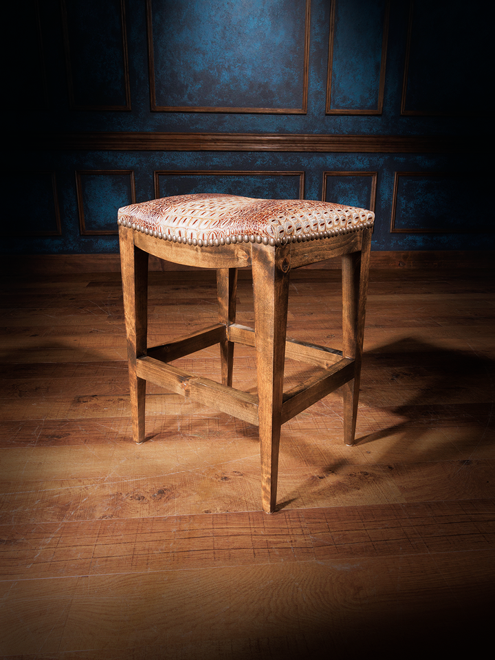 A brown crocodile leather saddle stool with a patterned fabric seat cushion, placed on a hardwood floor with a blue panelled wall in the background.