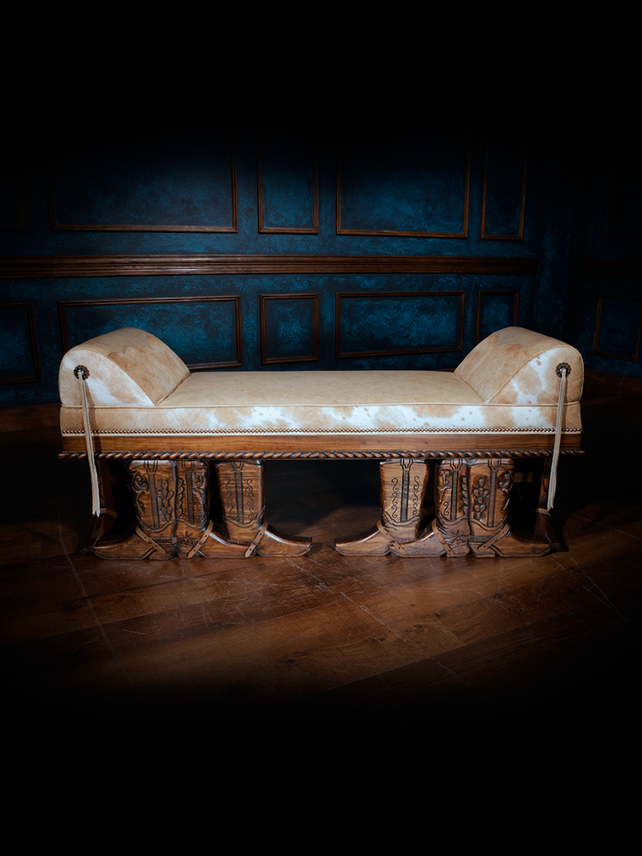 A bench with a blonde brindle cowhide seat and hand carved cowboy boot trim, set against a blue-painted wall with wooden flooring.