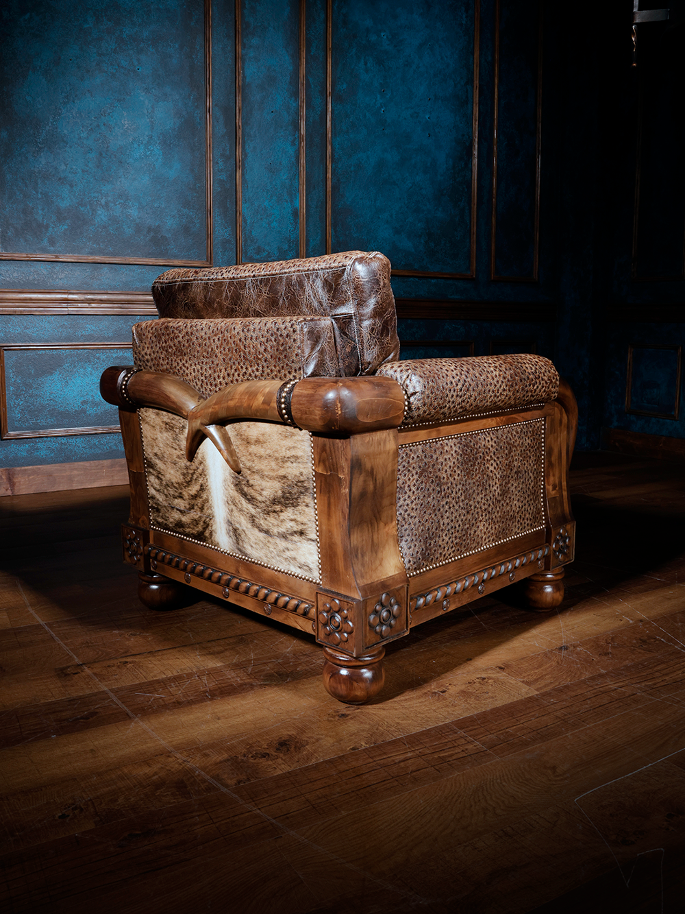 Rear and side angle of a western armchair showcasing tan and cream hair-on-hide panels, tooled leather upholstery, and rounded wood feet. The sculptural wood arms curve over the frame, blending craftsmanship with cowboy character.