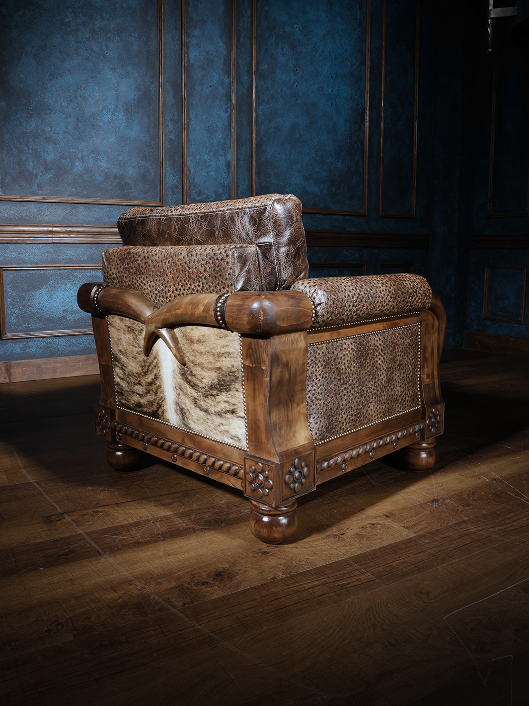Rear and side angle of a western armchair showcasing tan and cream hair-on-hide panels, tooled leather upholstery, and rounded wood feet. The sculptural wood arms curve over the frame, blending craftsmanship with cowboy character.