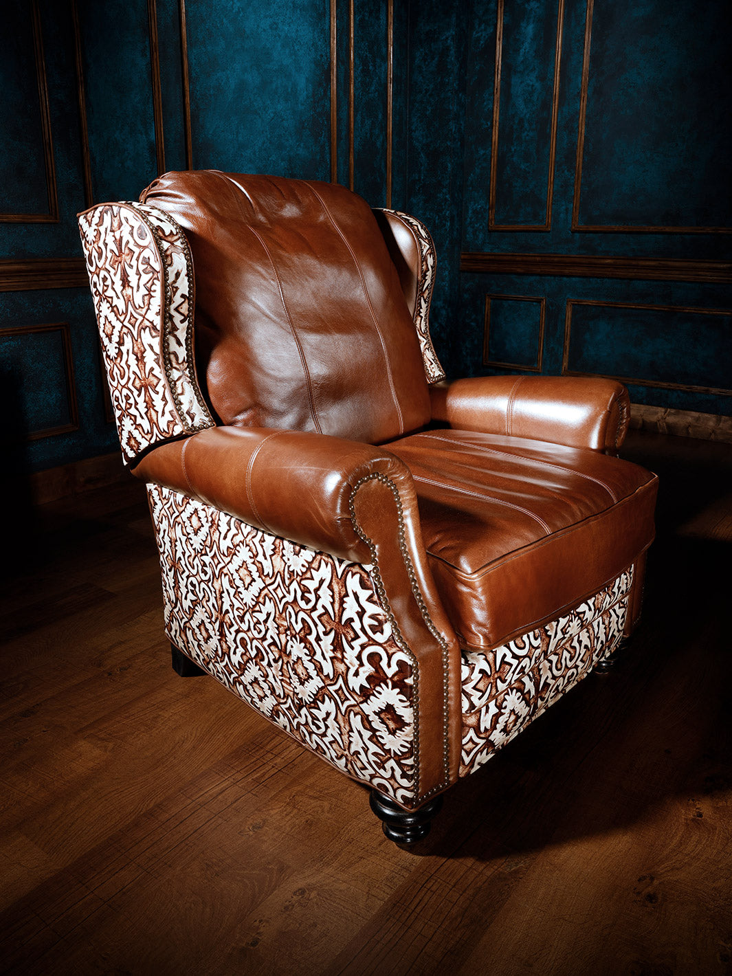 A luxurious leather recliner chair with a patterned backrest and a rich brown leather seat and armrests, set against a textured blue wall with ornate wood trim.