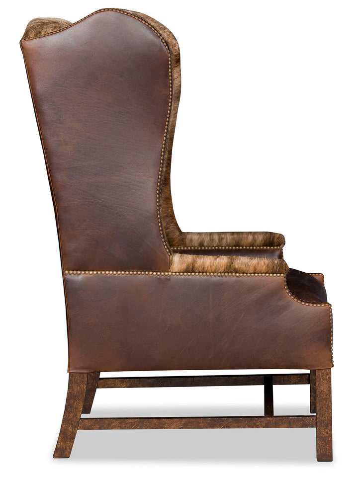 Side profile of the accent chair showing the elegant curve of the wingback, leather upholstery, and nailhead accents along the edges, supported by a distressed wood frame.
