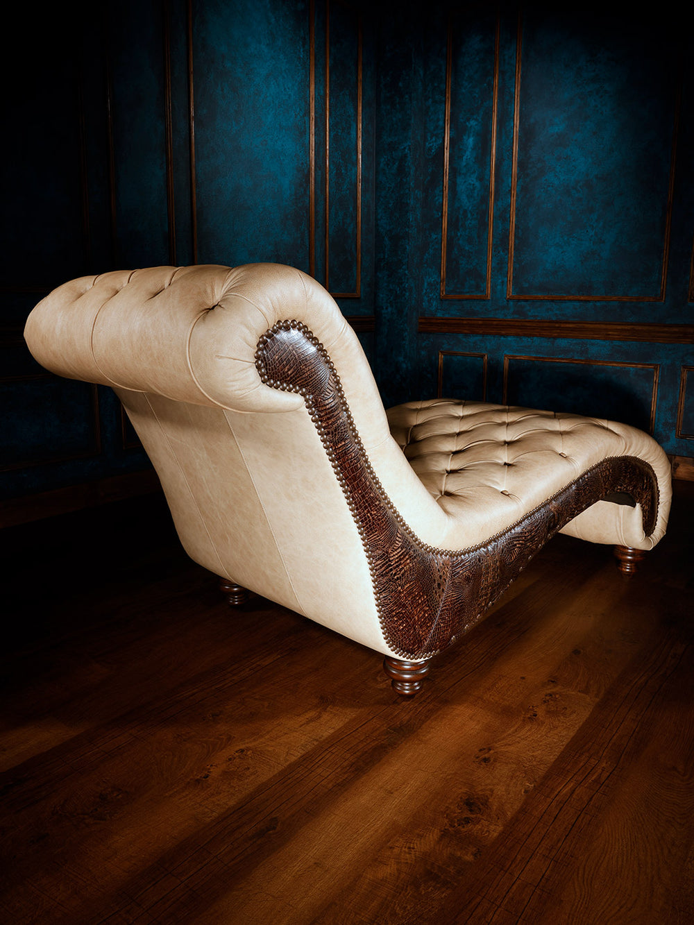 A side view of a luxurious chaise lounge showcasing its plush beige leather and intricate wooden craftsmanship, set in a dark, opulent room. The piece combines comfort with refined style.