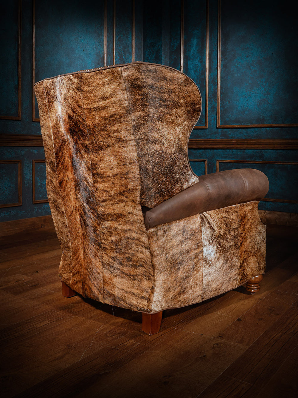 A western-style recliner featuring a cowhide-patterned back and sides with brown leather armrests, set in a room with dark blue walls and wood flooring.