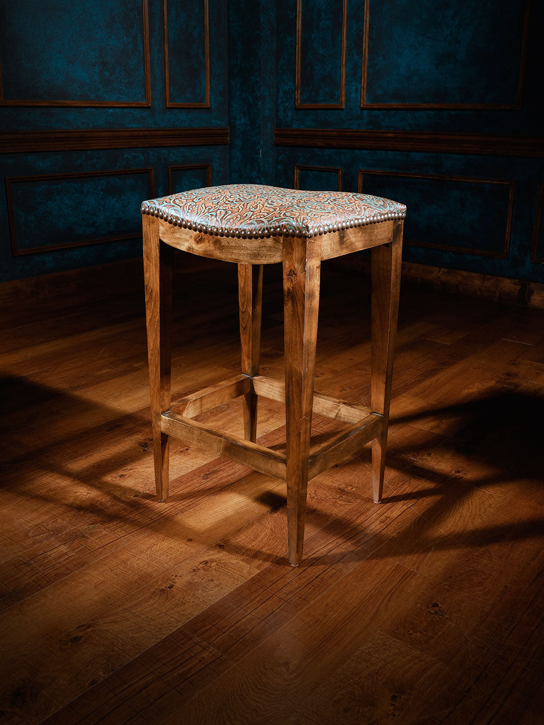A tooled leather saddle stool with a patterned top, placed on a hardwood floor against a blue wall.