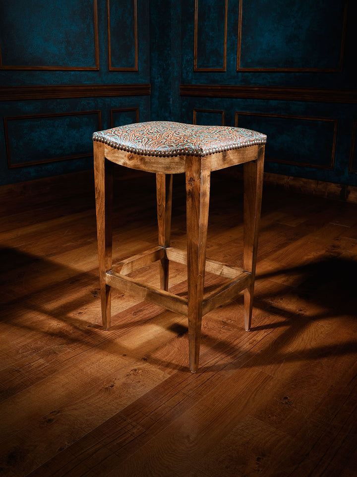 A tooled leather saddle stool with a patterned top, placed on a hardwood floor against a blue wall.