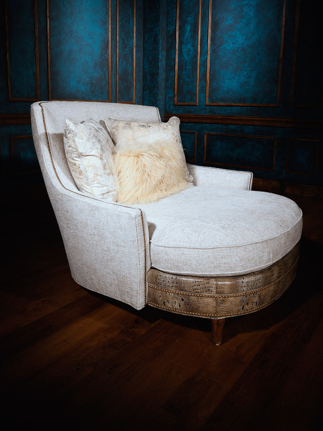 An ivory fabric chaise lounge with a cushion and a crocodile embossed accent, set against a blue panelled wall.