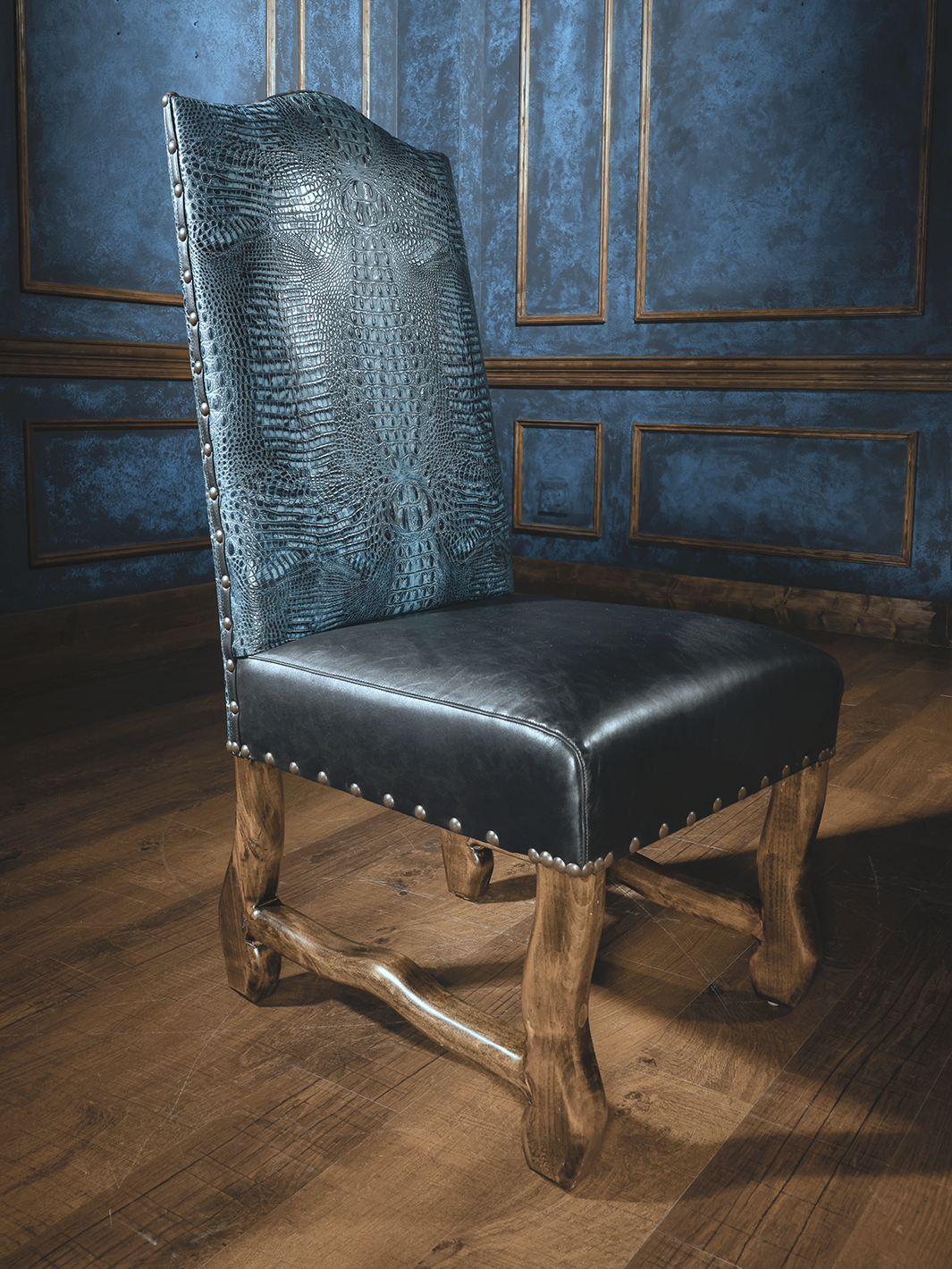 A royal blue leather dining chair with an embossed crocodile pattern, featuring a dark chocolate leather strap and wooden legs, positioned against a blue paneled wall.
