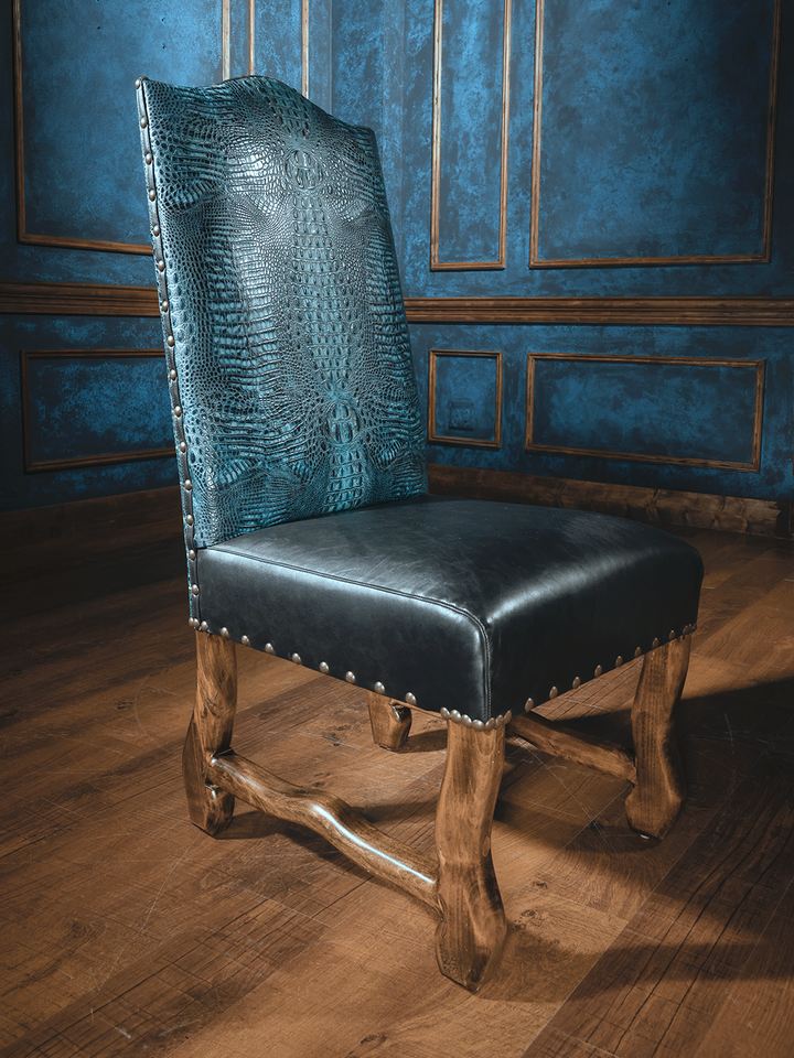 A royal blue leather dining chair with an embossed crocodile pattern, featuring a dark chocolate leather strap and wooden legs, positioned against a blue paneled wall.