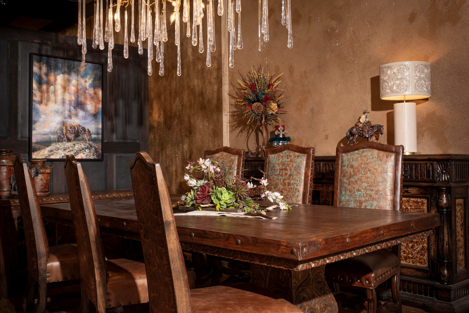 Western dining room with carved wood table, patina leather chairs, glass-drop chandelier, rustic sideboard, floral centerpiece, and buffalo wall art.