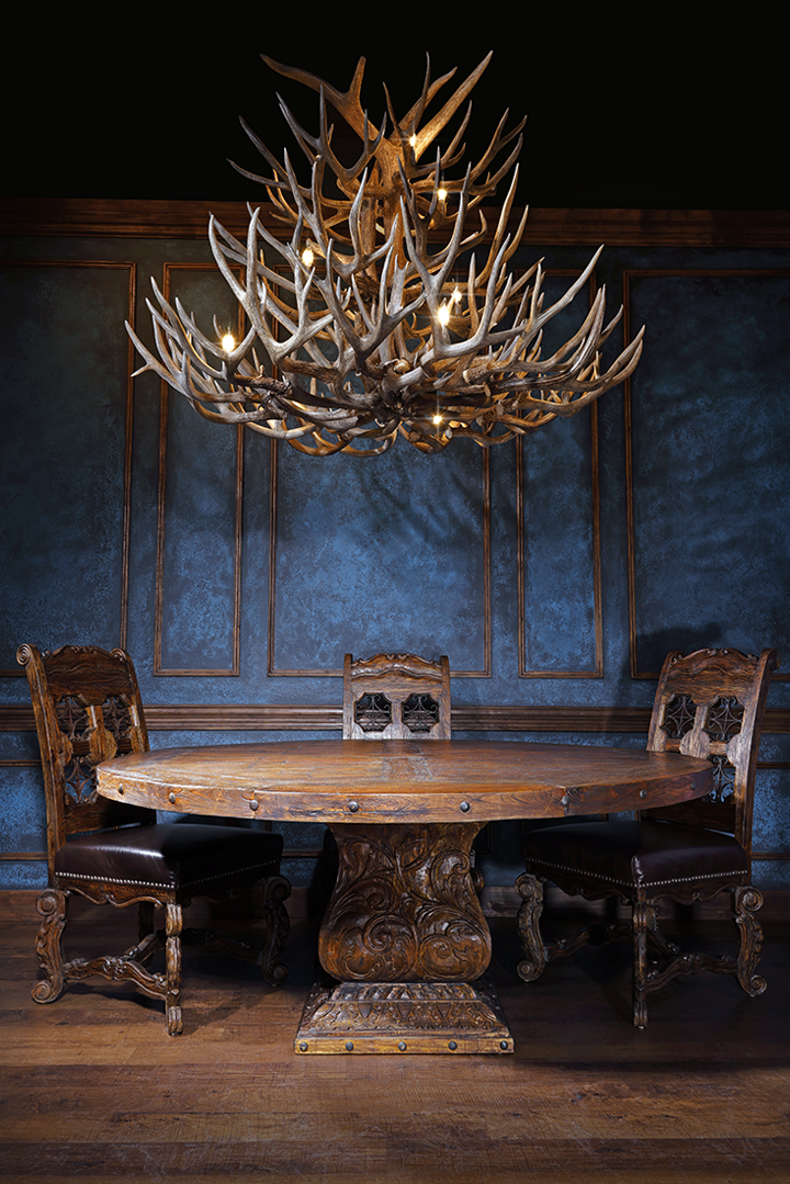 Western-style dining room featuring a hand-carved round wood pedestal table, leather dining chairs with star cutouts, and a large authentic antler chandelier against a deep blue paneled wall.