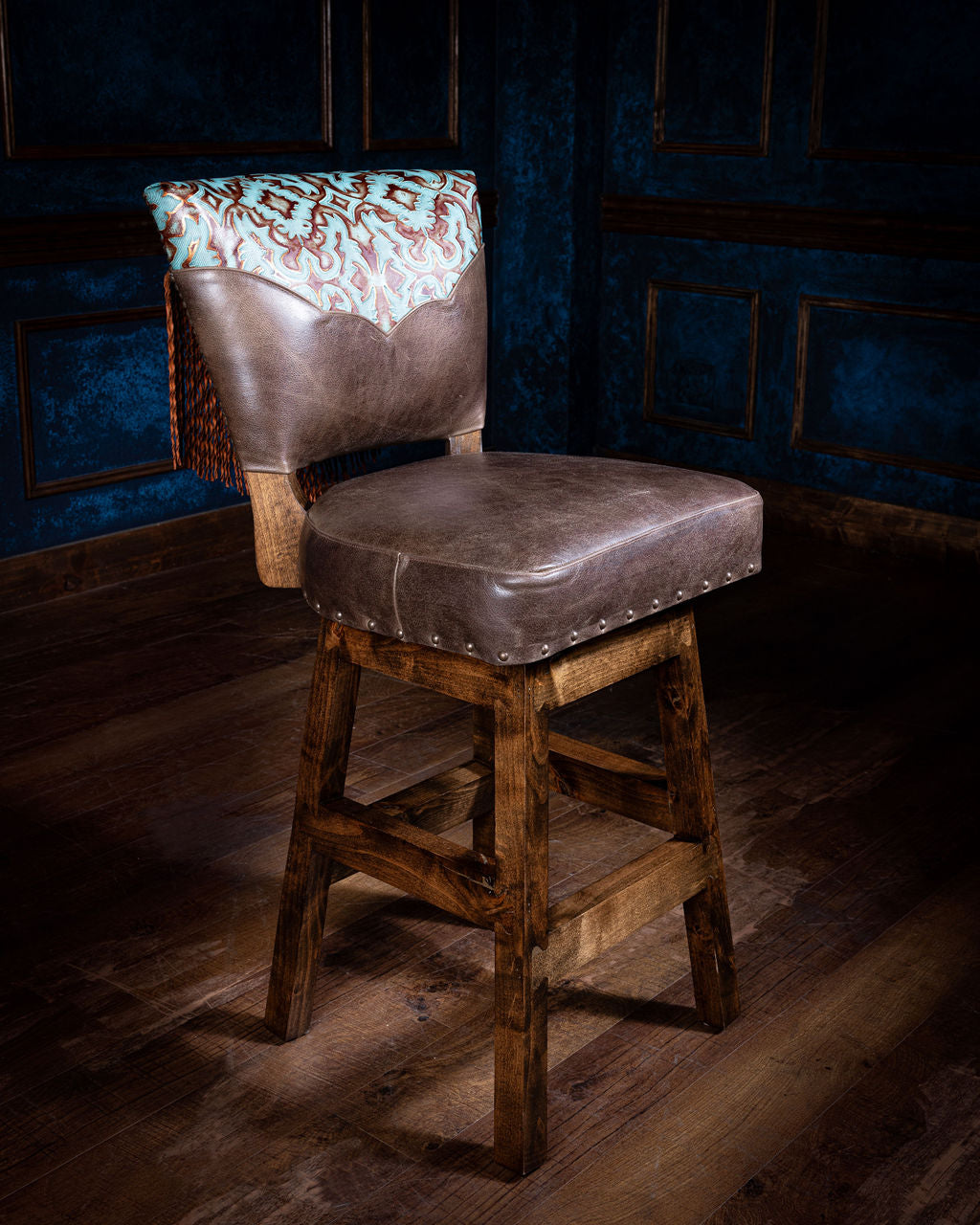 A brown wolf leather yoke bar stool with a decorative cushion on the top, set against an dark blue background with wooden flooring.