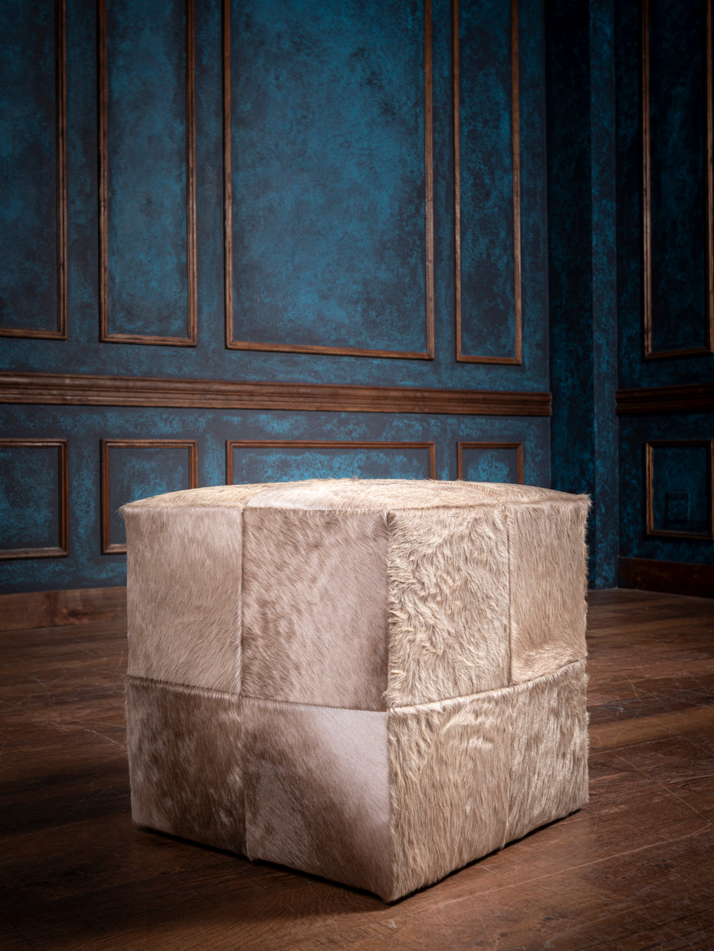 Cube-shaped ottoman covered in beige hair on hide fur, set against a teal-paneled wall with brown trim and wooden floor.